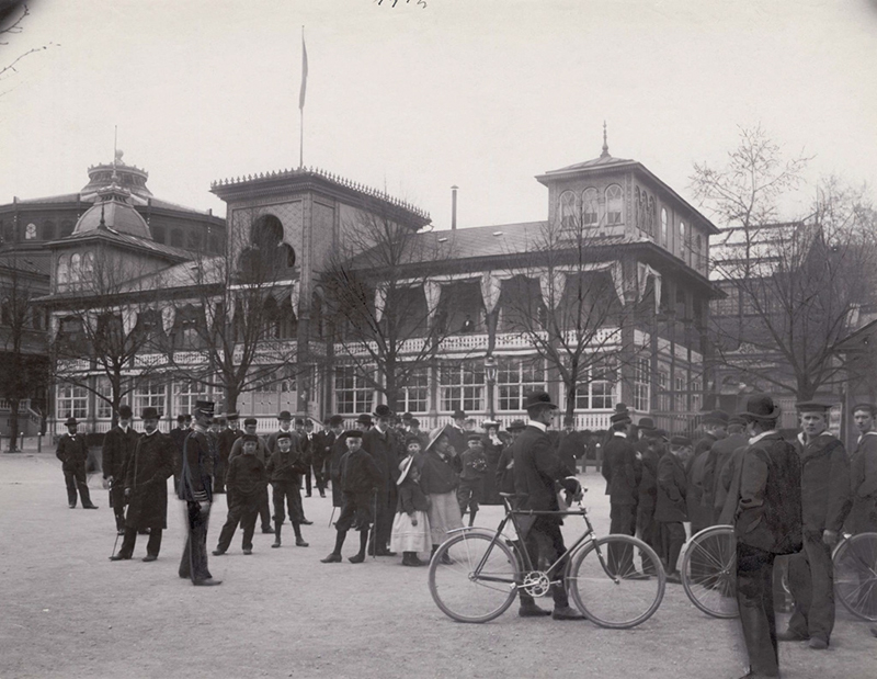 Alhambra, restaurang och teater, på Djurgårdsslätten, Djurgården, Stockholm. Uppfört av hovkonditor Pohl omkring 1830. Även kallat Pohlsro. Byggnadens fasad i bakgrunden. En folksamling - män, barn, k